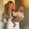 A postpartum mother in a white textured top and shorts holds a baby and a tin our Pure Collagen, standing against a warm-lit wall.