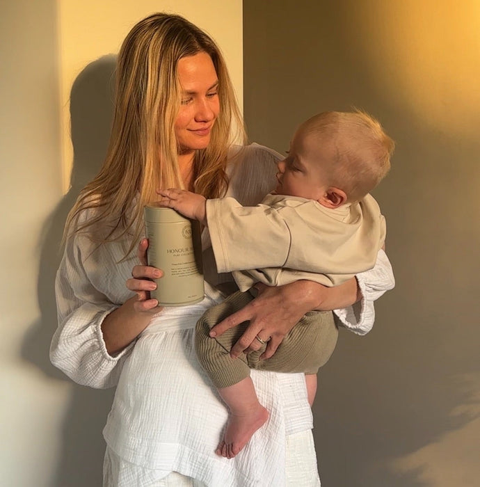 A postpartum mother in a white textured top and shorts holds a baby and a tin our Pure Collagen, standing against a warm-lit wall.
